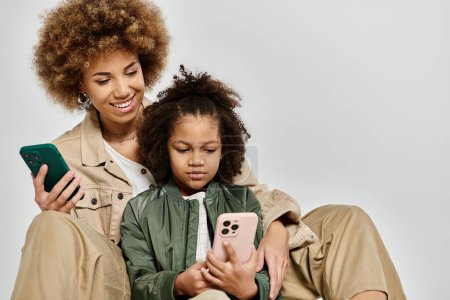 Curly African American mother and daughter in stylish clothes using cell phones while seated on a grey background.
