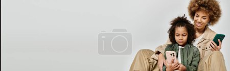 Photo for Curly African American mother and daughter, stylishly dressed, holding and looking at a cell phone on a grey background. - Royalty Free Image
