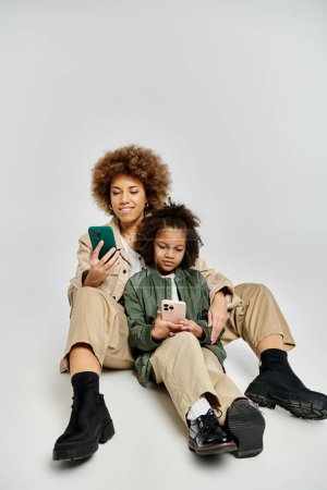 Una madre y una hija afroamericanas rizadas con ropa elegante sentadas en el suelo, profundamente absortas en usar un teléfono celular.