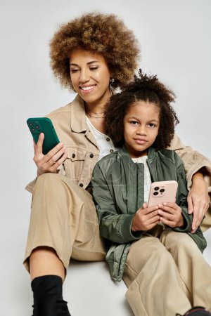 Lockige afroamerikanische Mutter und Tochter sitzen auf dem Boden, Mobiltelefone in der Hand, in stilvoller Kleidung auf grauem Hintergrund.