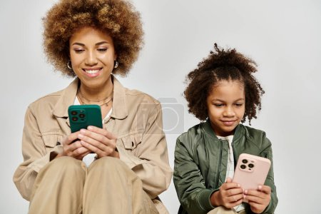 Una madre y una hija afroamericanas rizadas con ropa elegante enfocadas en sus teléfonos, absortas en mundos digitales.