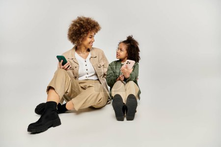 Lockige afroamerikanische Mutter und Tochter in stilvoller Kleidung auf dem Boden sitzend, in ein Handy vertieft.