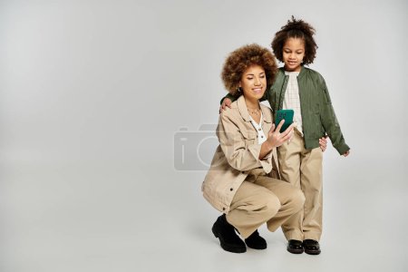 Una madre y una hija afroamericanas rizadas con ropa elegante posando juntas sobre un fondo gris.