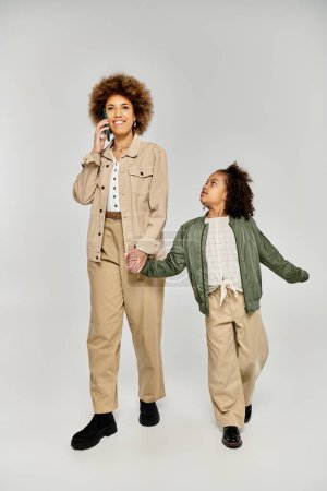 Lockige afroamerikanische Mutter und Tochter in stylischer Kleidung führen vor grauem Hintergrund ein Telefongespräch.