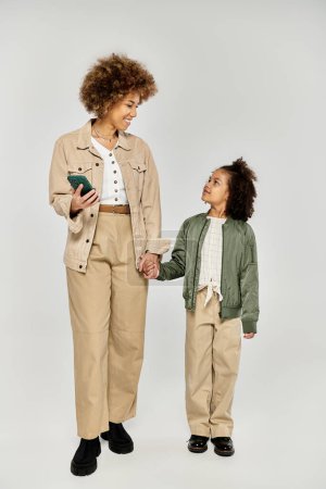 Lockige afroamerikanische Mutter und Tochter in stylischer Kleidung, Händchen haltend vor grauem Hintergrund.