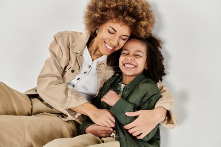 Eine lockige afroamerikanische Mutter umarmt ihre Tochter liebevoll auf dem Boden in stilvoller Kleidung vor grauem Hintergrund.