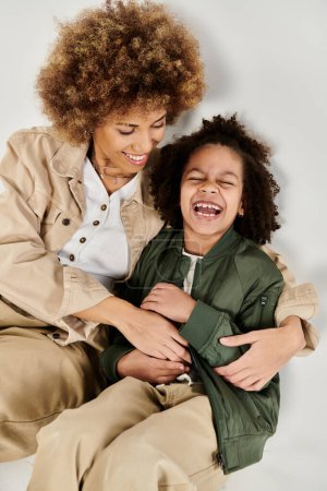 Eine lockige afroamerikanische Mutter und ihre Tochter lachen in stilvollen Kleidern auf dem Boden und schaffen gemeinsam glückliche Erinnerungen.