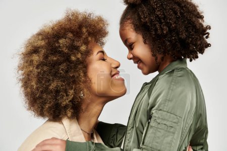 Une mère et une fille afro-américaine, toutes deux avec des cheveux bouclés, partageant un câlin affectueux sur fond gris.