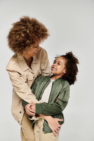 Foto de Una madre y una hija afroamericanas rizadas con ropa elegante se abrazan en un abrazo conmovedor contra un fondo blanco. - Imagen libre de derechos