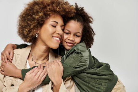 Foto de Madre e hija afroamericana rizada con ropa elegante abrazándose ferozmente frente a un fondo blanco. - Imagen libre de derechos