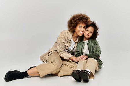 Curly African American mother and daughter in stylish attire, sitting on the floor, smiling warmly at each other.