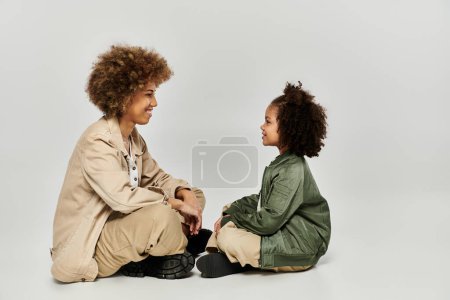 A curly African American mother and daughter sit closely on the floor, sharing a tender moment in stylish clothing.