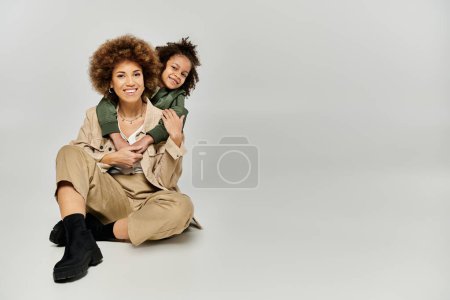 Foto de Una madre y una hija afroamericanas rizadas, ambas vestidas con estilo, se sientan juntas sobre un fondo gris. - Imagen libre de derechos