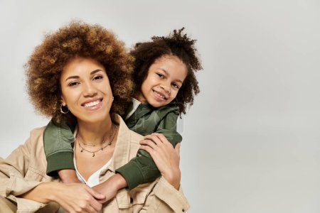 Stilvolle afroamerikanische Mutter mit lockigem Afrohaar trägt ihre Tochter auf den Schultern vor grauem Hintergrund.