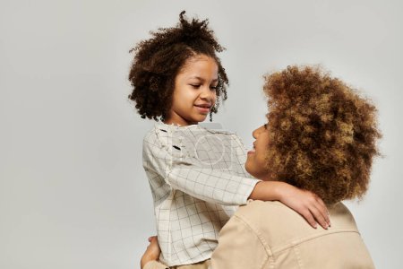 Foto de A curly African American mother and daughter wearing stylish clothes hug each other affectionately on a gray background. - Imagen libre de derechos