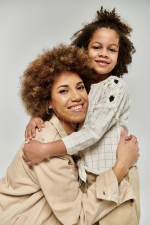 Foto de Madre e hija afroamericana rizada con ropa elegante abrazándose amorosamente contra un fondo gris. - Imagen libre de derechos