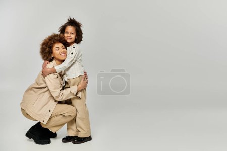 Foto de Una madre afroamericana rizada abraza calurosamente a su hija, ambas vestidas con estilo, sobre un fondo gris sereno. - Imagen libre de derechos
