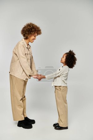 Téléchargez les photos : Une mère et une fille afro-américaines frisées, vêtues de vêtements élégants, se tiennent côte à côte sur un fond gris. - en image libre de droit