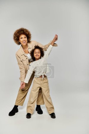 Téléchargez les photos : Une mère afro-américaine frisée et sa fille dans des vêtements élégants frappant une pose sur un fond gris. - en image libre de droit