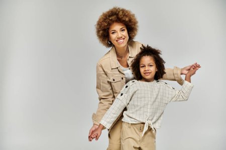 A curly African American mother and daughter, in stylish clothing, striking a pose on a grey background.