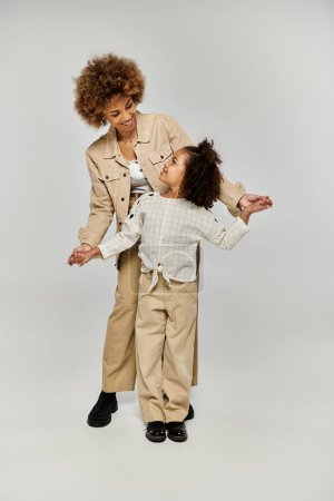 Une mère et une fille afro-américaines frisées se tiennent avec confiance devant un fond blanc, mettant en valeur leurs tenues élégantes.
