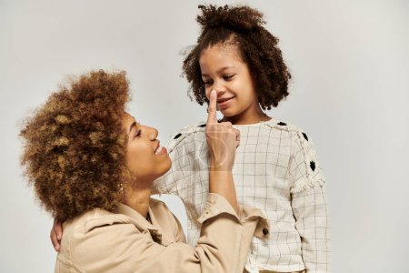Photo for A curly African American mother touching nose of daughter in stylish attire against a grey backdrop. - Royalty Free Image