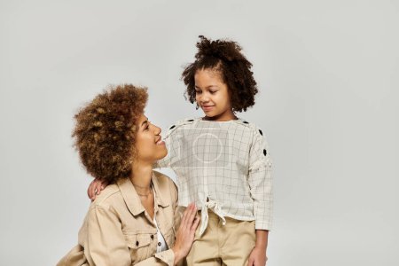 Foto de Una madre y una hija afroamericanas rizadas con ropa elegante posando elegantemente sobre un fondo gris. - Imagen libre de derechos