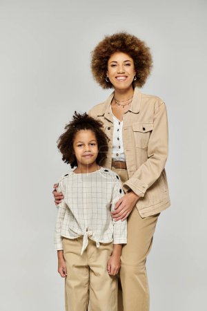 La mère et la fille afro-américaines aux cheveux bouclés prennent une pose élégante sur fond gris neutre.