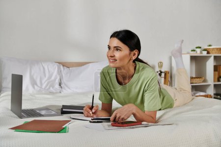 Woman in casual attire relaxing on bed, writing in notebook.