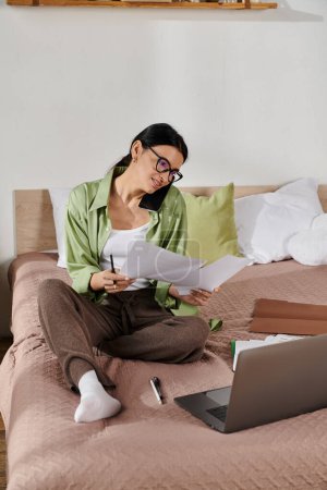 A woman in casual attire works on her laptop surrounded by papers on a cozy bed.