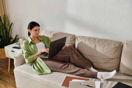 A casually dressed woman uses a laptop on a couch.