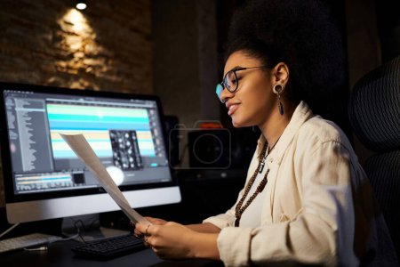 A woman with glasses sits at a computer in a recording studio, focused and engaged in her digital work.