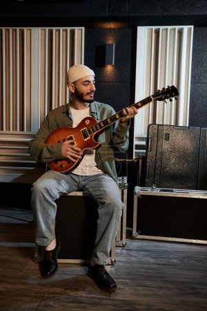 Un músico sentado en un estudio de grabación rasga una guitarra, profundamente inmerso en la creación de música para las bandas próxima actuación.