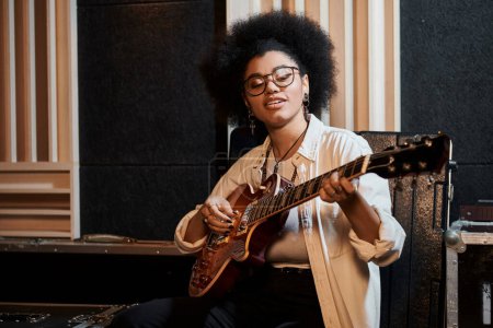 A woman in glasses playing a guitar with passion in a recording studio during a music band rehearsal.