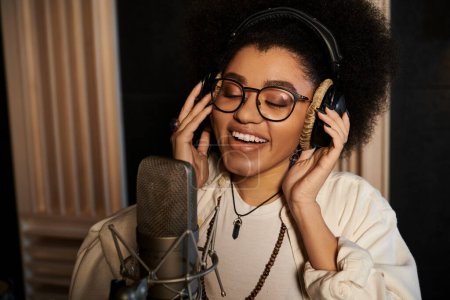 Une femme aux lunettes et aux cheveux afro chante passionnément dans un microphone lors d'une répétition d'un groupe de musique dans un studio d'enregistrement.