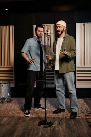 Two men stand before a microphone in a recording studio during a music band rehearsal.