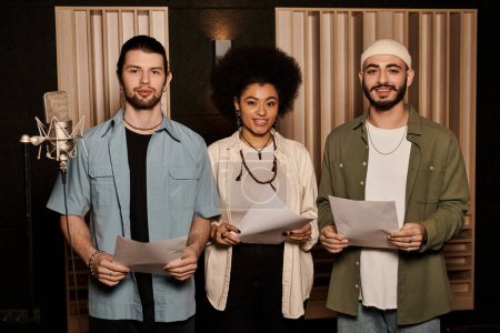 Three individuals stand in a recording studio holding lyric sheets, preparing for a music band rehearsal session.
