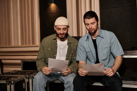 Two musicians with papers sit on a bench, engrossed in composing music for their bands rehearsal in a recording studio.