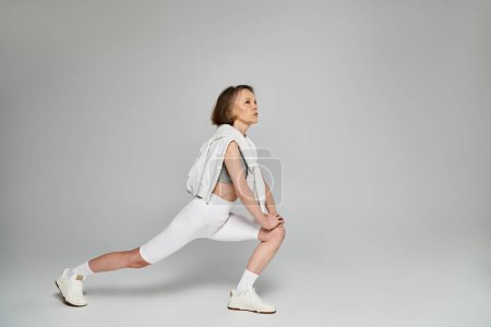 Mature woman in comfortable attire performing a squat on a white background.