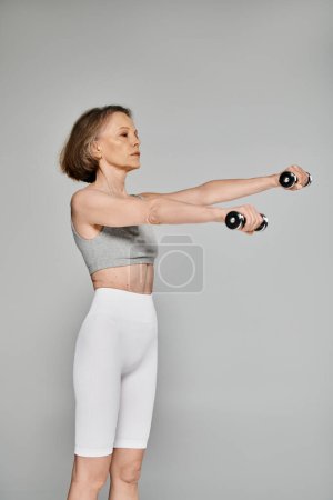 Attraktive Frau in aktiver Kleidung beim Training mit Kurzhanteln auf grauem Hintergrund.