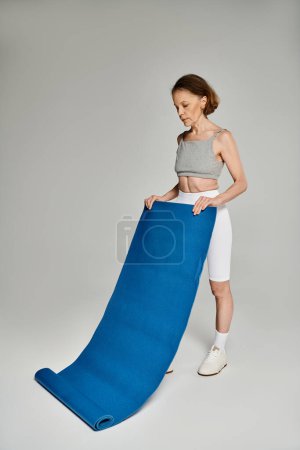 A mature woman in comfortable clothing holding a large blue yoga mat.