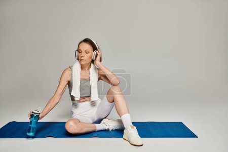 Reife Frau in bequemer Kleidung sitzt auf Yogamatte und hört Musik.