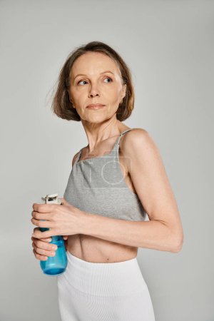 Active woman in comfy attire holding a water bottle on gray background.