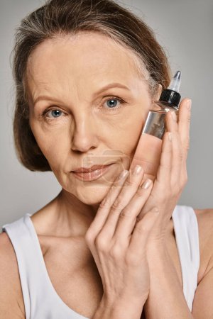 Photo for A mature, attractive woman in cozy attire holds a bottle of facial serum. - Royalty Free Image