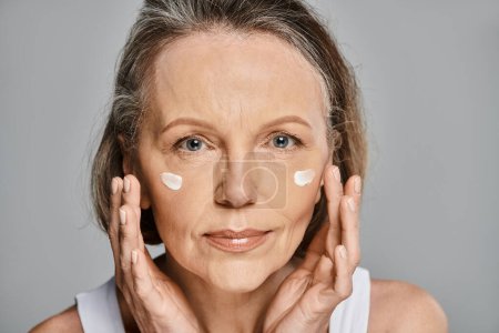 An older woman serenely applying cream to her face.