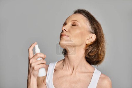 A mature woman in comfy attire is actively using a spray on her face for a refreshing moment.