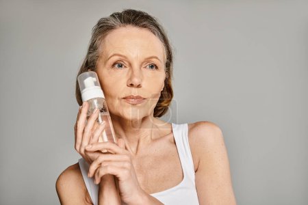 A mature woman in cozy attire holding a facial cleanser bottle.