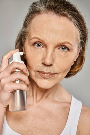 A mature, attractive woman in comfy attire holds a bottle of skin care product.