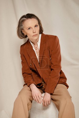 Stilvolle reife Frau in braunem Blazer und brauner Hose sitzt bequem auf einem Hocker.
