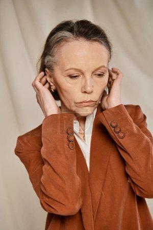 Mature woman in tan jacket poses gracefully with hands on ears.